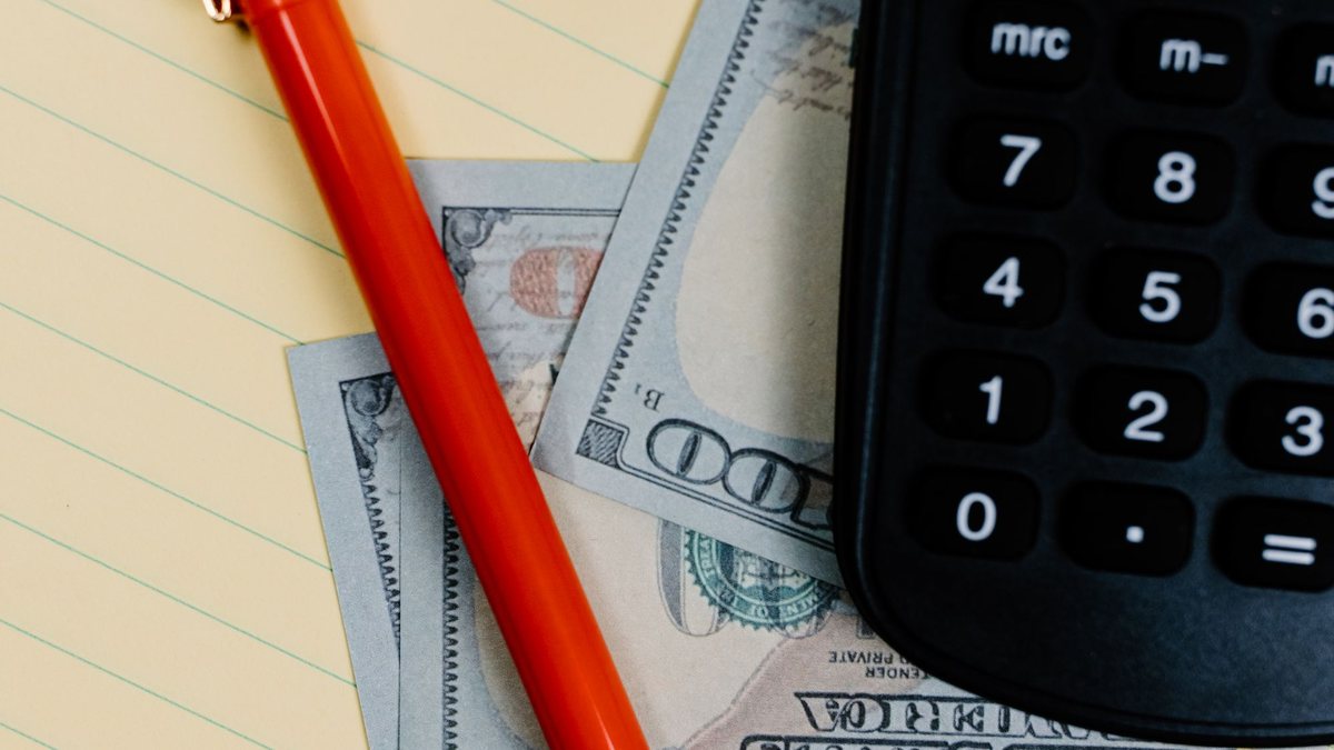 Red pen, dollar bills and a calculator on a notepad for pricing creative work