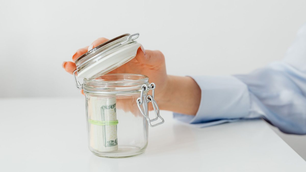 Hand holding a small glass jar with rolled-up dollar bills inside