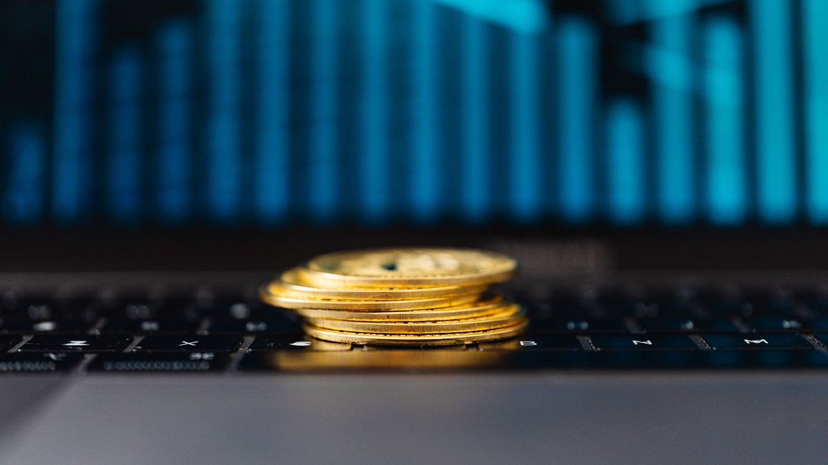 Stack of gold coins on a laptop keyboard in front of a glowing bar chart