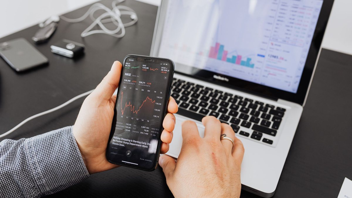 Hands holding a smartphone with a stock chart in front of a laptop showing market data