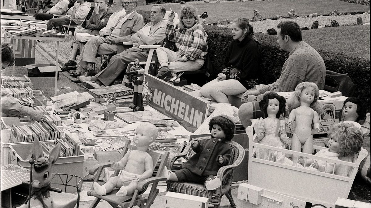Black-and-white photo of a busy neighborhood garage sale with tables of toys and household goods