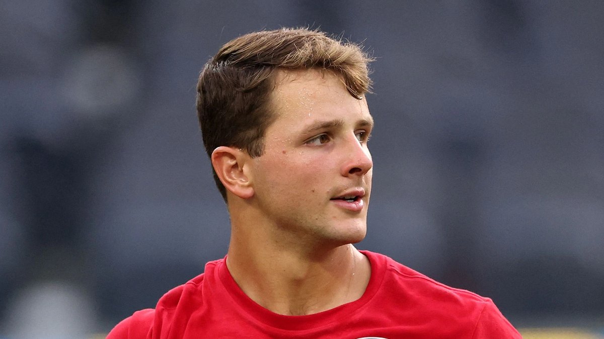 49ers quarterback Brock Purdy in a red 49ers warmup shirt holds a football before a preseason game in 2026