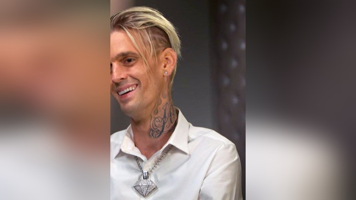 Aaron Carter smiling in a white shirt and a diamond pendant during an interview