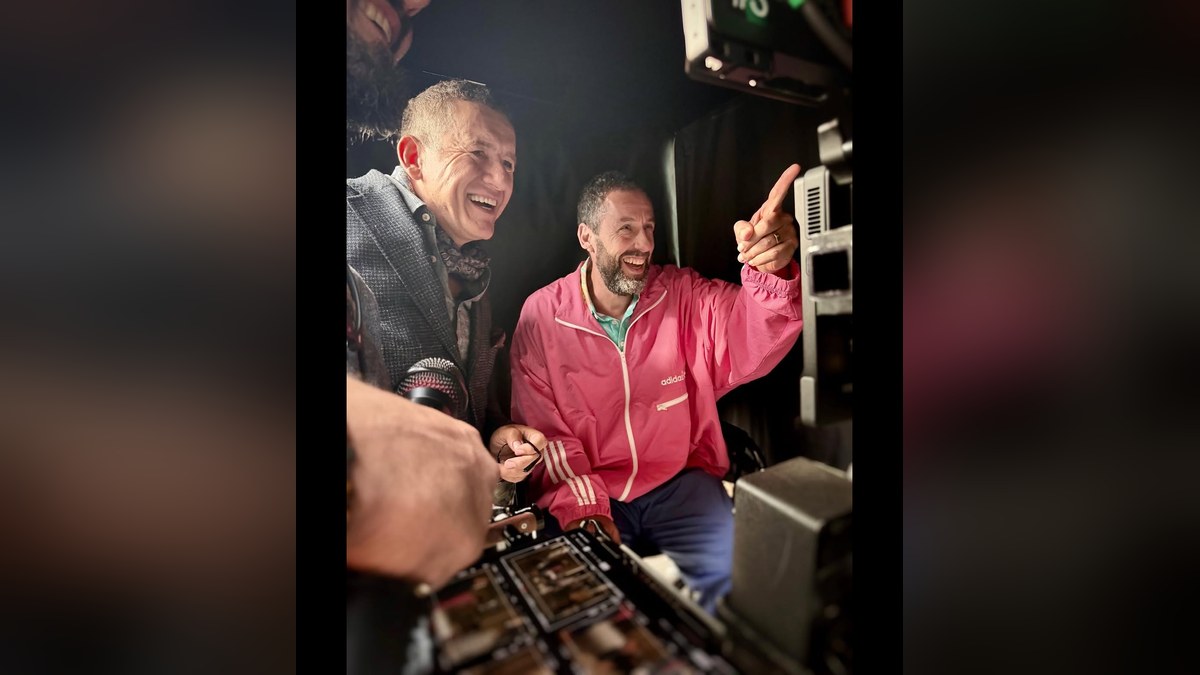 Adam Sandler in a pink jacket laughing and pointing beside Dany Boon on a film set