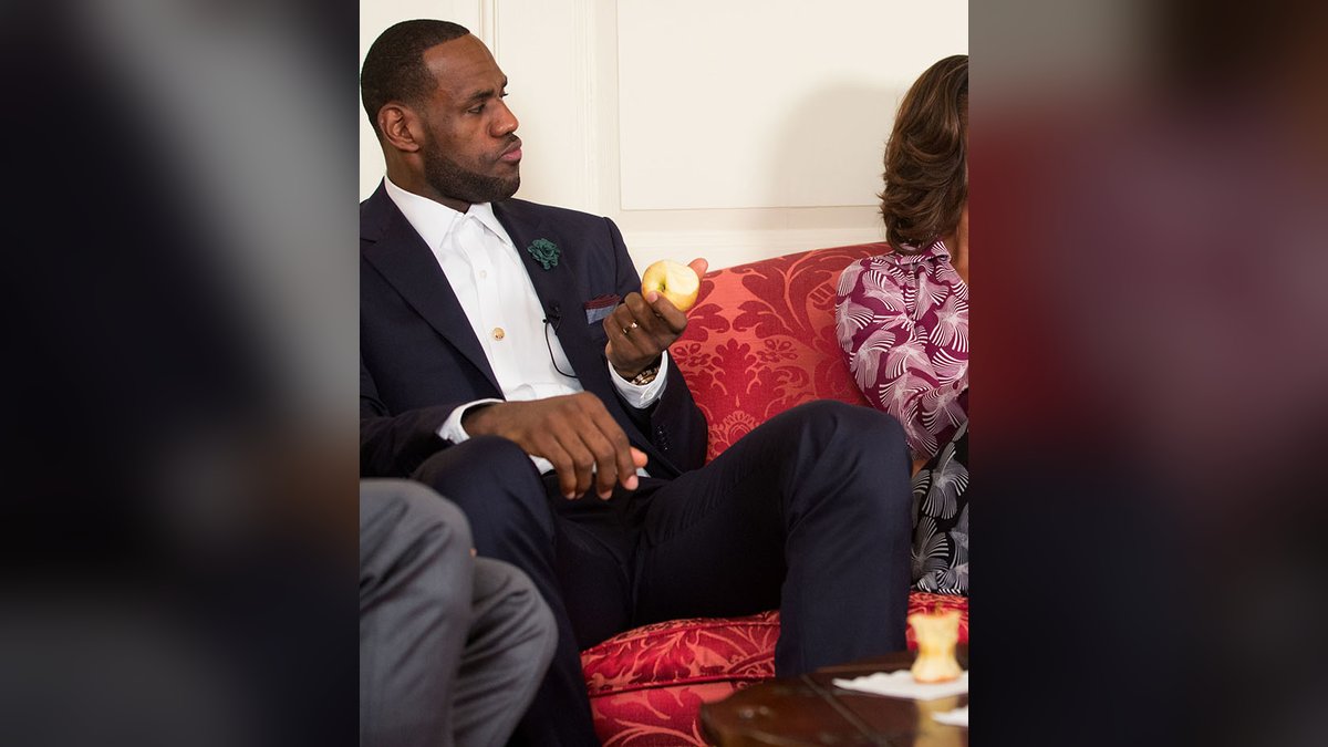 LeBron James in a dark suit holds an apple while seated in the Map Room of the White House in 2014