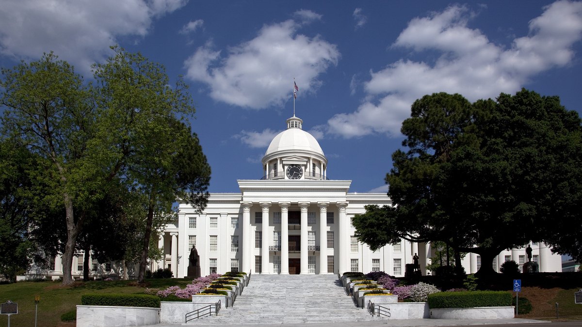 The white columned Alabama State Capitol with its dome rises behind a wide stairway lined with flower beds and leafy oak trees