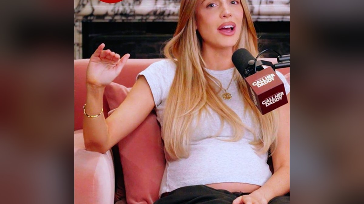 Alex Cooper talking in a white T-shirt as she shows her baby bump on a pink sofa