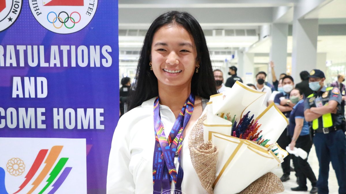 Alex Eala smiles in a white jacket with a medal lanyard around her neck while holding a bouquet of flowers on her arrival in Manila in 2022