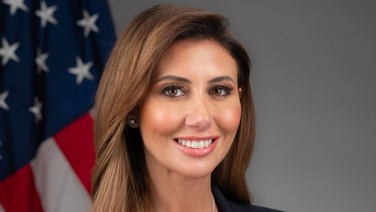 Alina Habba smiling in a navy pinstripe suit in front of an American flag