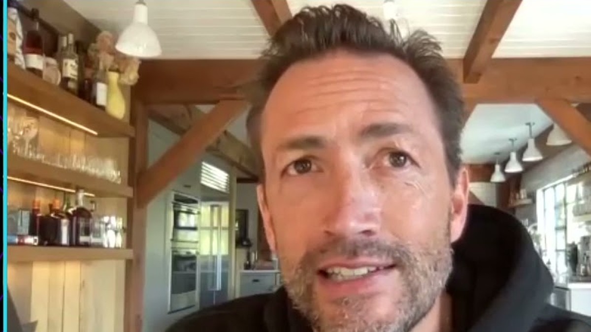 Andrew Shue in a black hoodie speaking on a video call from a sunny kitchen