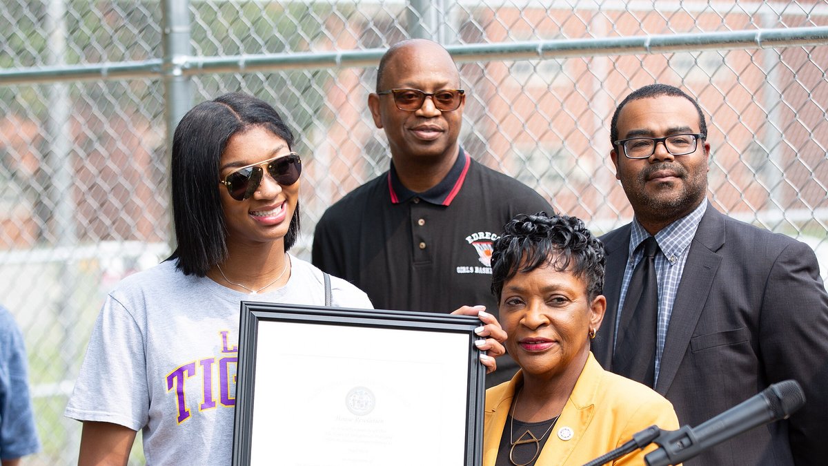 Angel Reese smiling in sunglasses as she holds a framed proclamation at the dedication of a basketball court named in her honor in Randallstown, Maryland