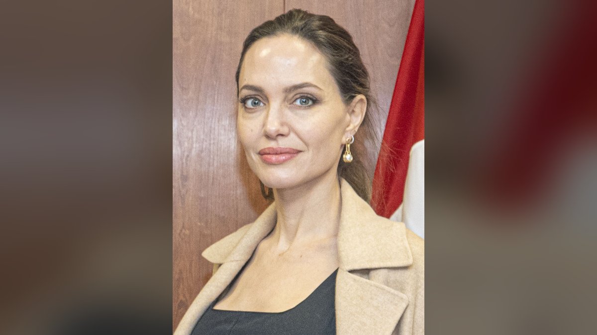 Angelina Jolie in a camel coat in front of a wood-paneled wall