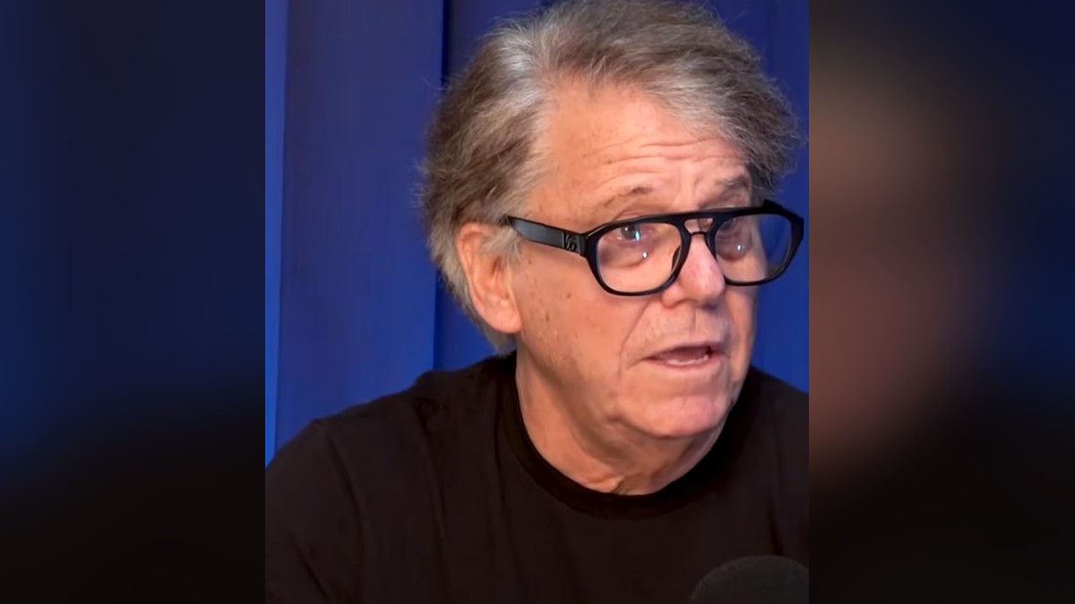 Anson Williams in black-framed glasses and a black T-shirt talking in front of a blue curtain
