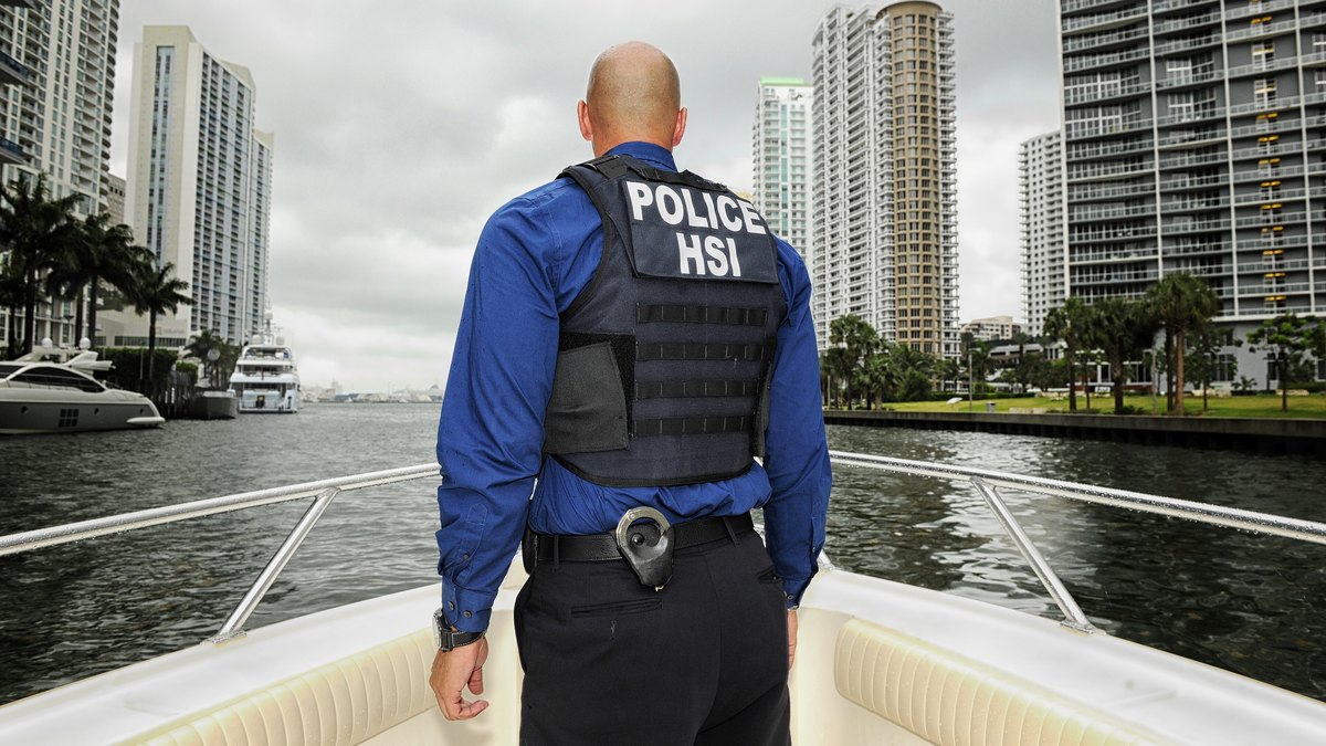 A federal agent in a blue shirt and a black vest marked Police HSI stands on the bow of a boat facing city high-rises