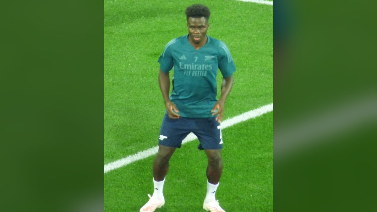 Arsenal winger Bukayo Saka in a teal Arsenal training top and navy shorts warming up on the field before a Champions League match
