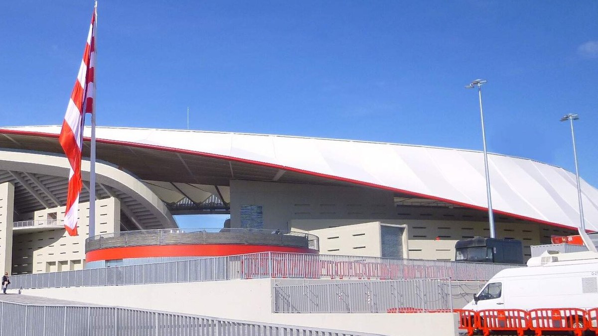 The curved white roof and red trim of Atlético Madrid's stadium under a clear blue sky with a red and white striped flag out front