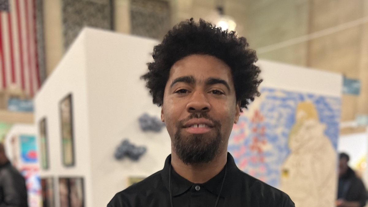 Baltimore photographer Devin Allen smiling at an art fair in front of a gallery wall of paintings