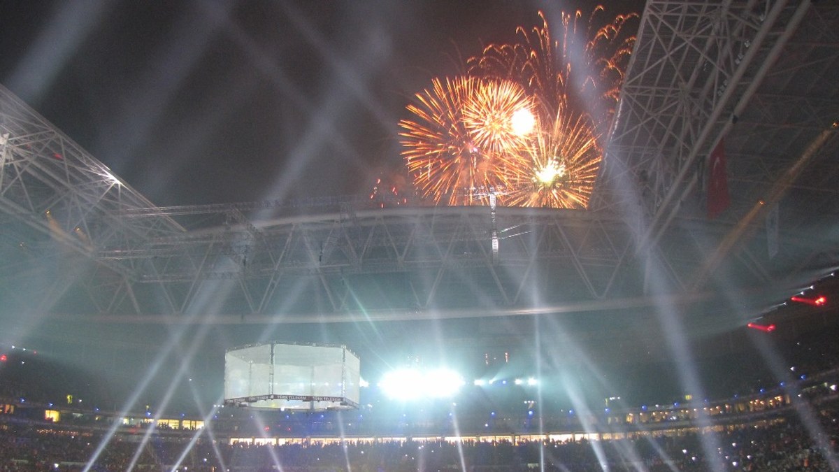 Fireworks and bright floodlights fill a packed soccer stadium under an open roof at night