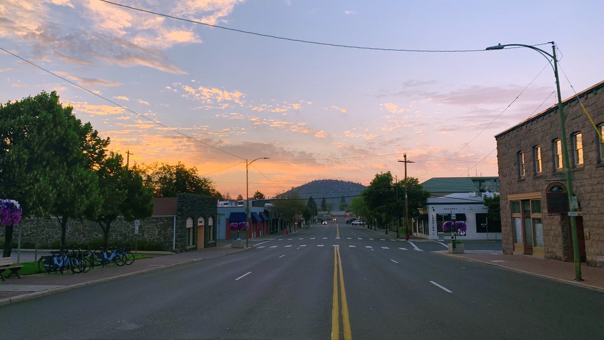 An empty downtown street in Bend, Oregon, at sunrise with stone buildings, hanging flower baskets and a pine-covered butte under a pink sky