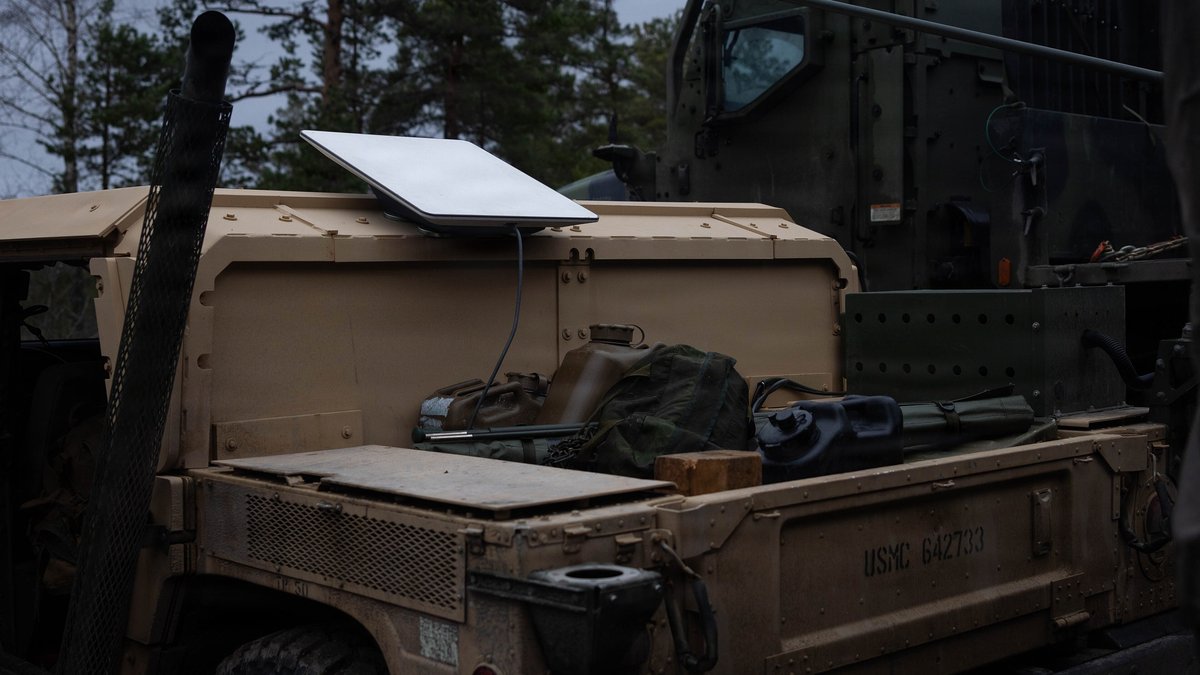 A flat white satellite internet dish sits on the back of a tan U.S. Marine Corps Humvee parked among pine trees