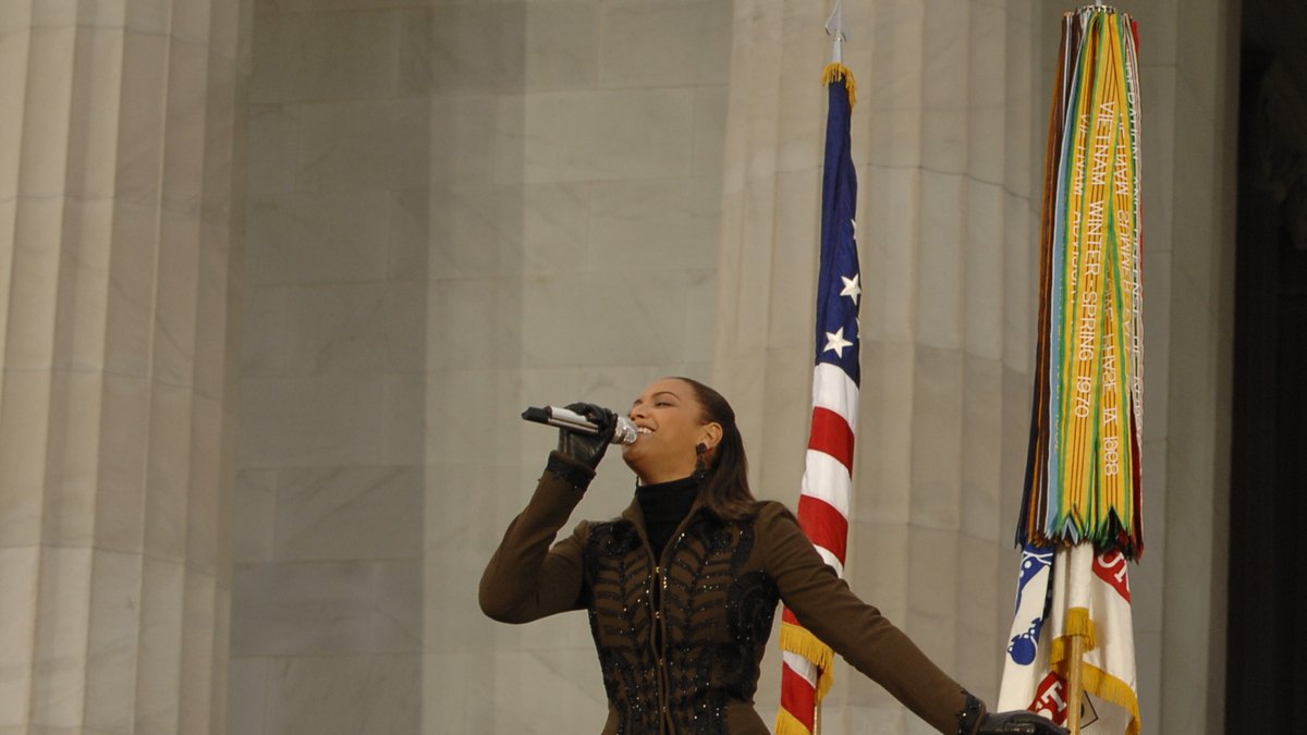 Beyoncé sings into a microphone in front of an American flag and military streamers at the Lincoln Memorial