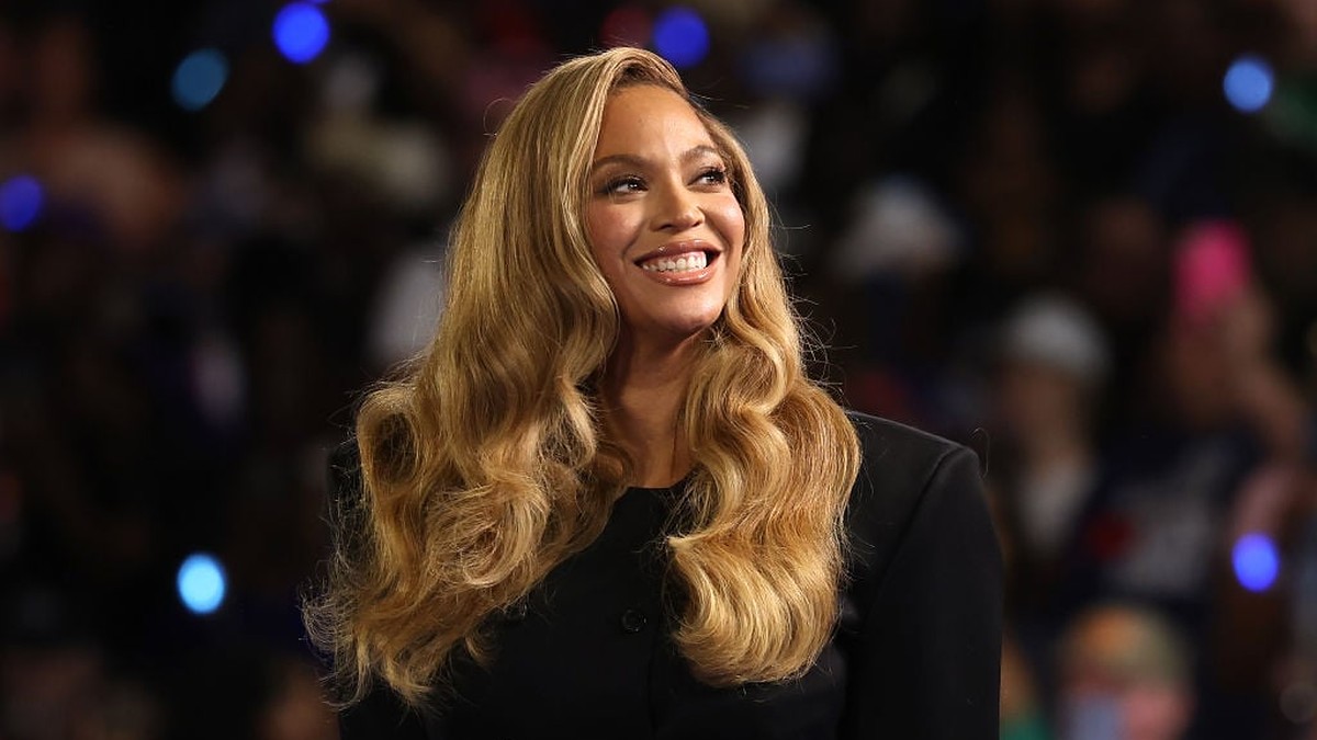 Beyoncé smiling with long honey-blonde waves in a black double-breasted blazer in front of a blurred crowd