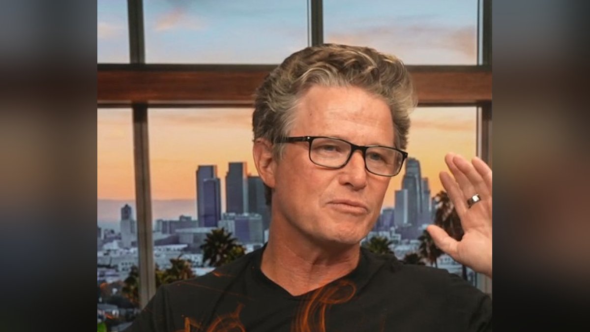 Billy Bush in glasses and a black T-shirt giving a small wave in front of a city skyline window
