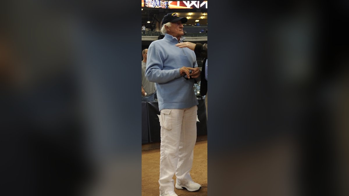 Bob Uecker in a light blue sweater and Brewers cap talks on the field at Miller Park before a 2011 Milwaukee Brewers game