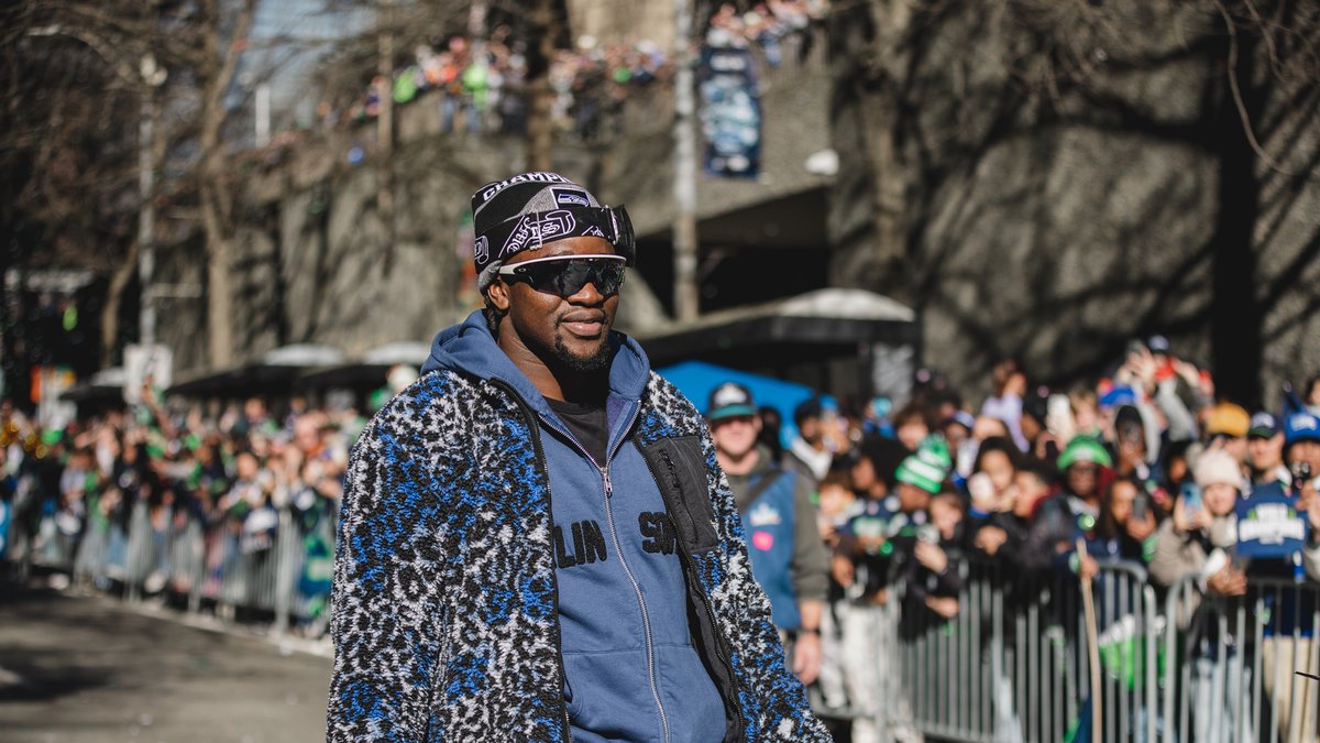 Boye Mafe smiling in sunglasses and a blue hoodie as fans line the street at the Seattle Super Bowl victory parade