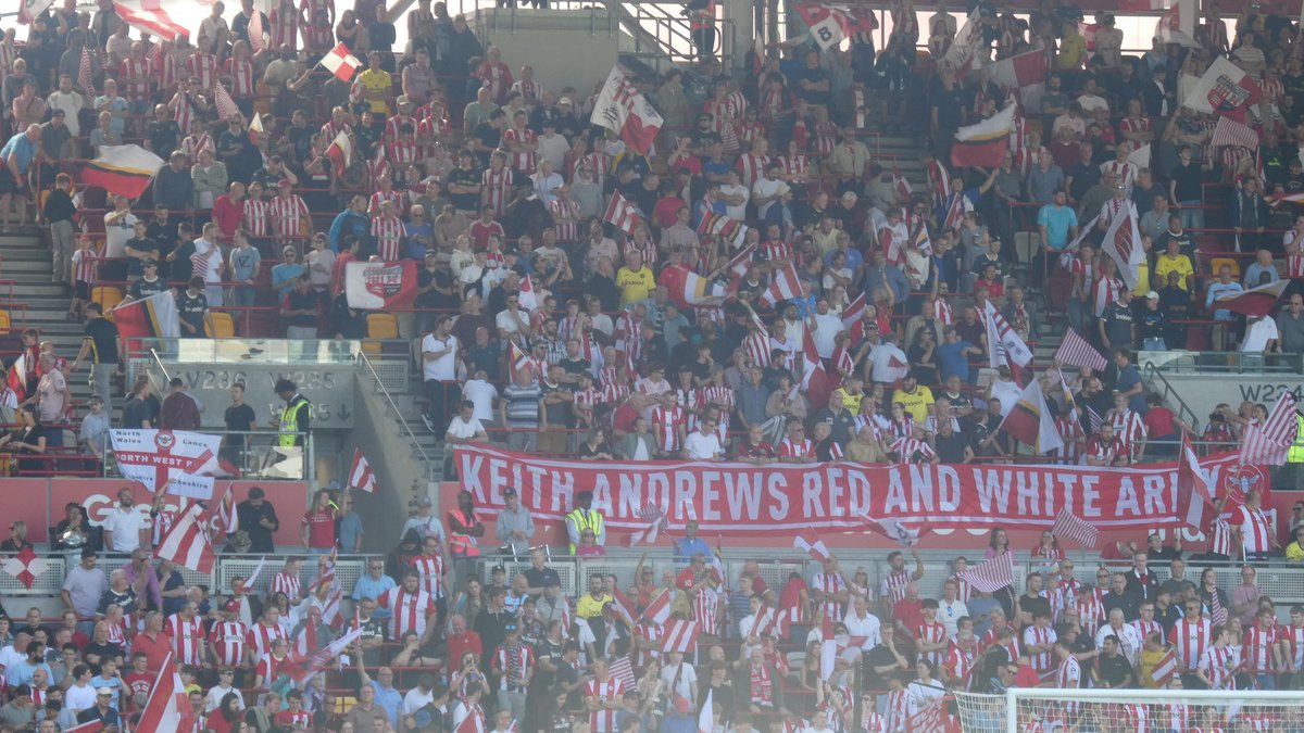 Brentford supporters in red and white shirts wave flags in the stands at the Gtech Community Stadium behind a banner for manager Keith Andrews