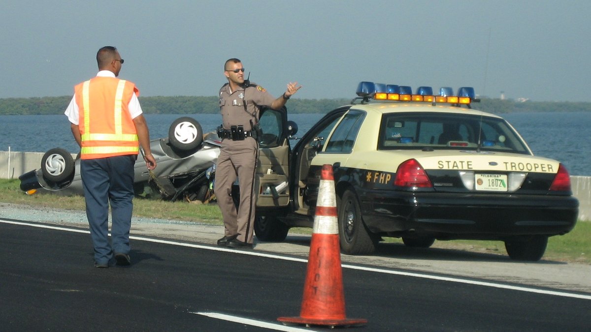 A Florida state trooper directs traffic beside his patrol car and an orange cone at a crash scene on a coastal highway