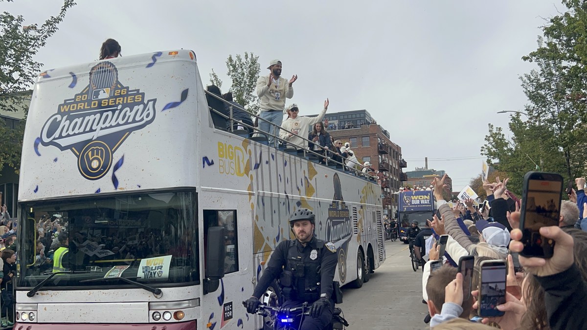 Milwaukee Brewers players wave from an open-top bus wrapped in 2026 World Series Champions graphics as fans line a downtown street