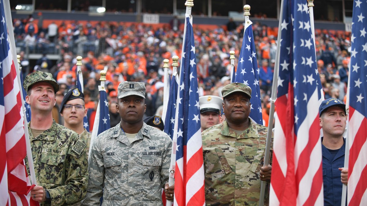 Service members hold American flags on the field in front of a crowd in orange at a Denver Broncos game