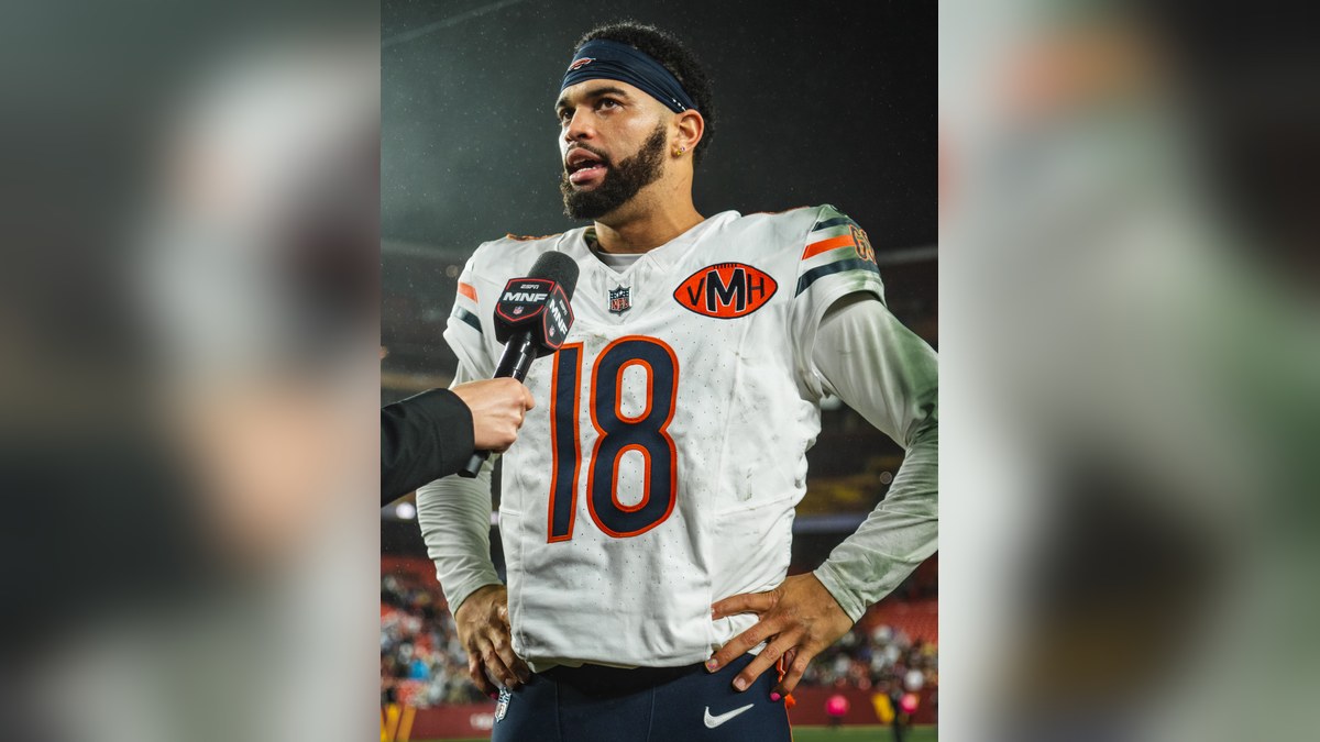 Caleb Williams in a white Chicago Bears No. 18 jersey talking with reporters on the field