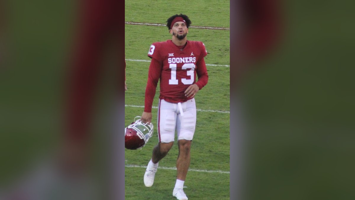 Caleb Williams in a red number 13 jersey walks on the field holding his helmet during a college game in 2021