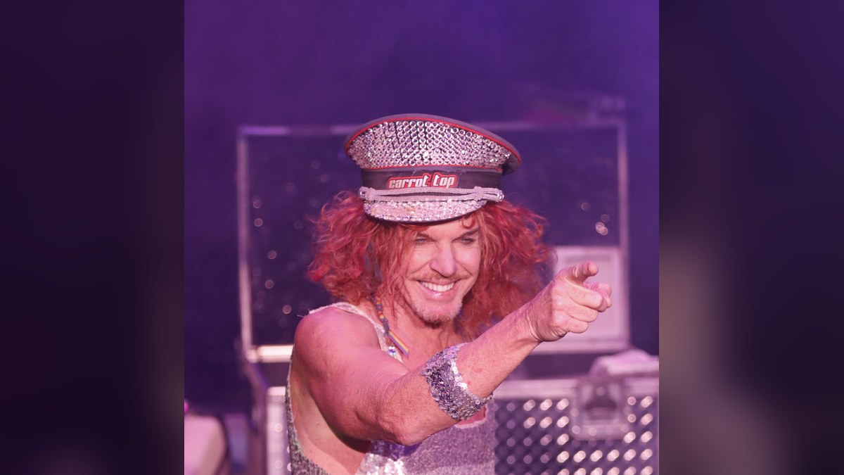 Carrot Top smiling and pointing on stage in a sequined top and jeweled cap