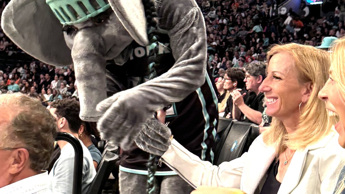 WNBA commissioner Cathy Engelbert smiles in the stands at Barclays Center as Ellie the Elephant, the New York Liberty mascot, leans in beside her