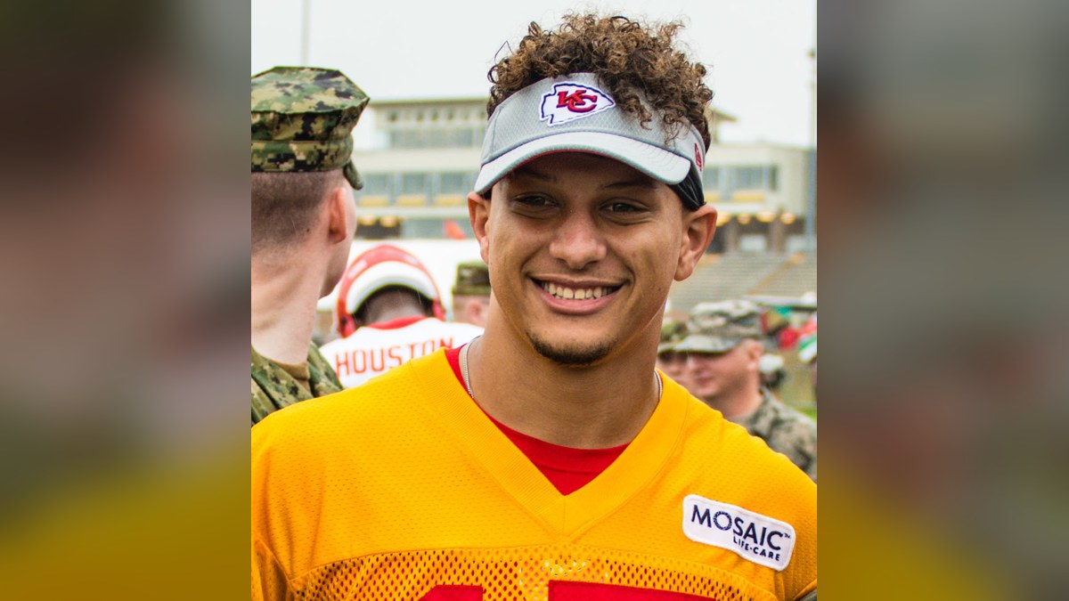 Patrick Mahomes smiling in a red Chiefs visor and yellow practice jersey at Kansas City Chiefs training camp