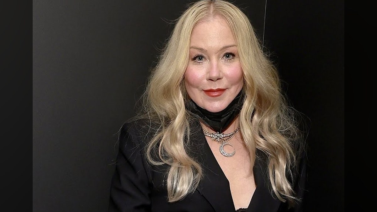 Christina Applegate with long wavy blond hair in a black blazer and crescent moon necklace