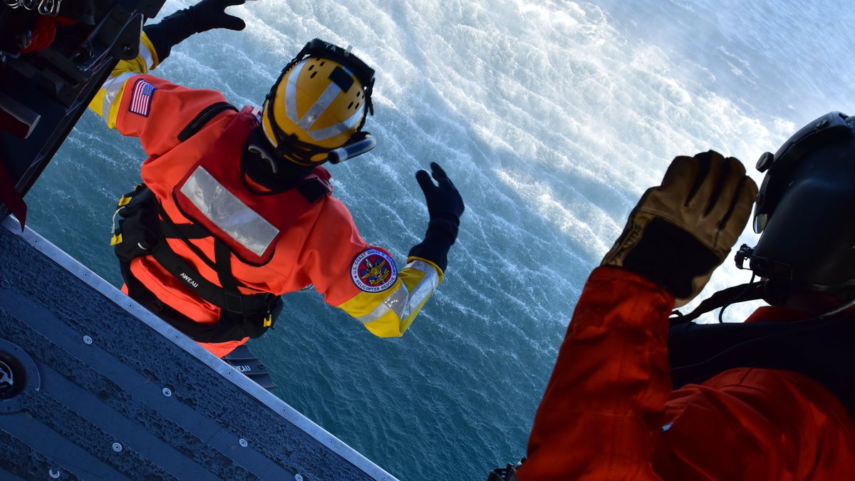 A U.S. Coast Guard rescue swimmer in an orange suit and yellow helmet leans out of a helicopter door high above the ocean