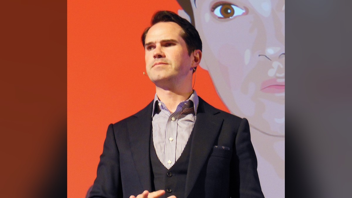Jimmy Carr in a dark three-piece suit gesturing as he speaks on a stage