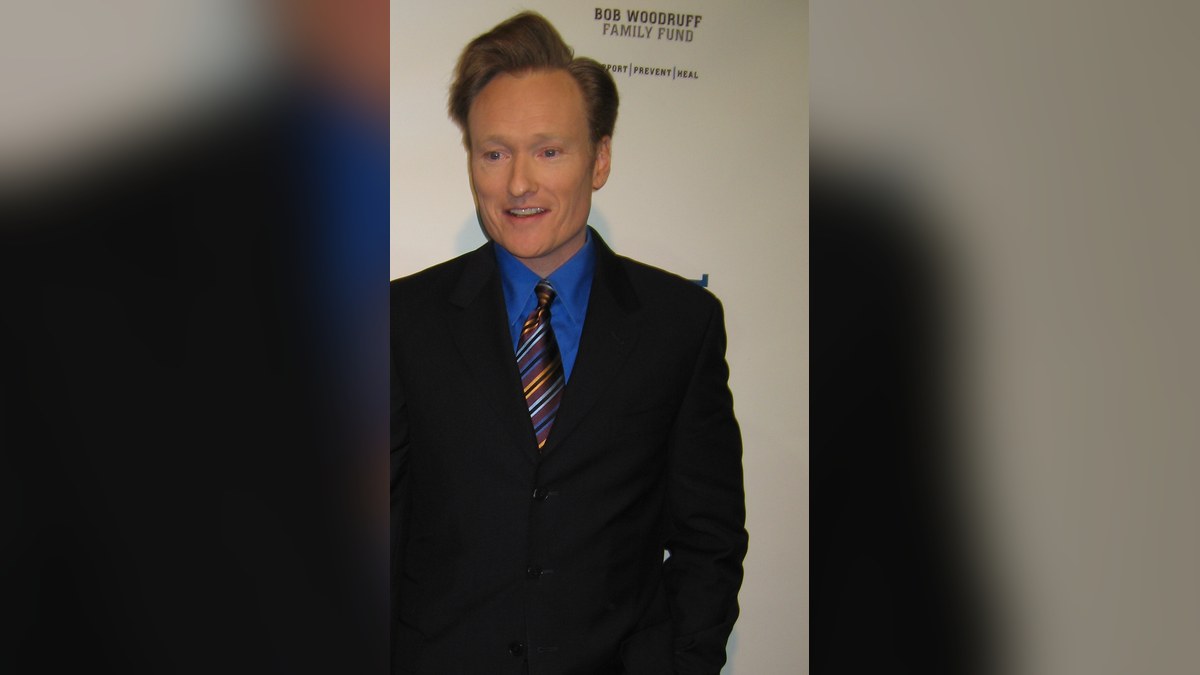 Conan O'Brien smiles in a dark suit and striped tie at a charity comedy benefit