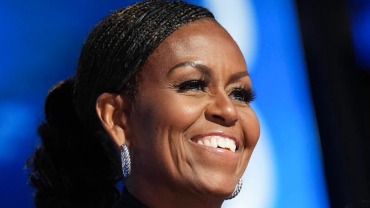 Michelle Obama smiling in silver hoop earrings in front of a bright blue stage backdrop