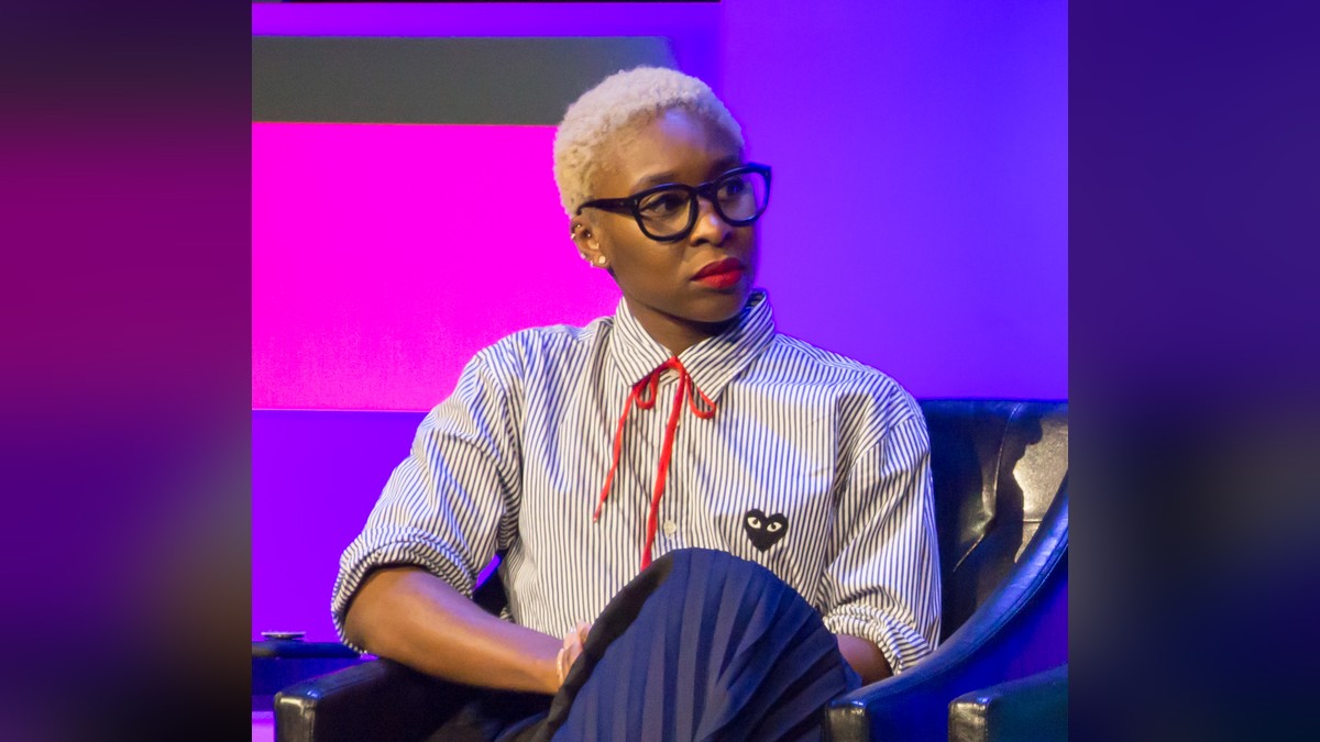 Cynthia Erivo in black glasses and a striped shirt with a red bow tie seated on stage at a panel