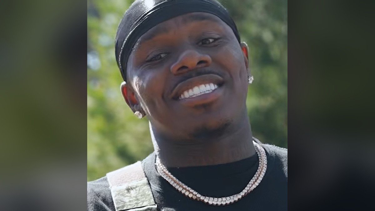 DaBaby smiling outdoors in a black cap and diamond chain