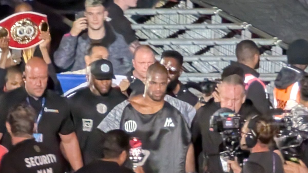Daniel Dubois in a gray shirt walks toward the ring through security staff and cameramen as a fan holds up a championship belt behind him