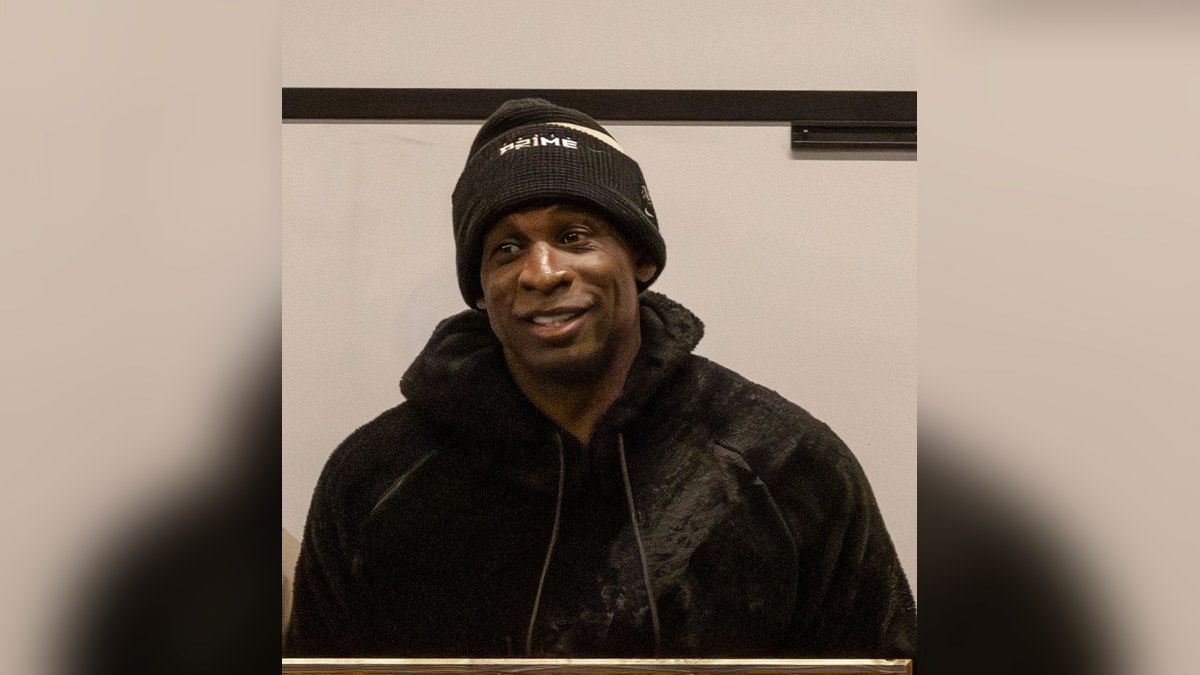 Deion Sanders smiling in a black Prime beanie and a black fleece hoodie at an event in Boulder