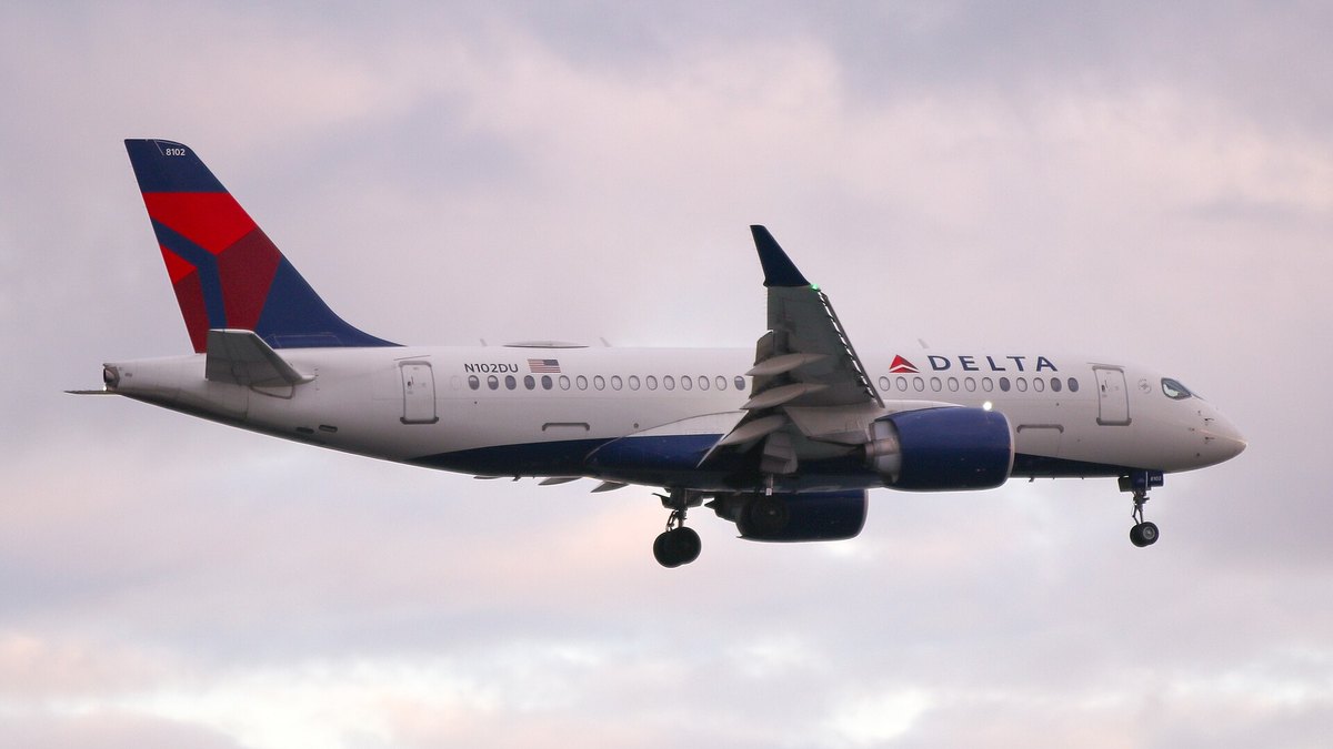 A red white and blue Delta passenger jet flies low with its landing gear down against a soft pink sky