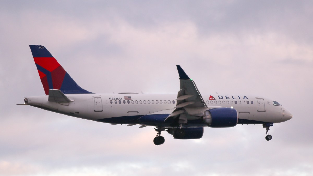 A white Delta Air Lines jet with a red and blue tail flying low under a pink cloudy sky