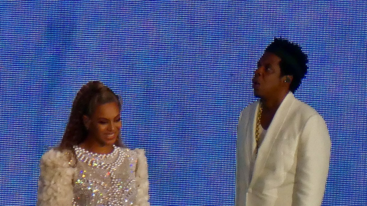Beyoncé smiling in a sparkling outfit beside Jay-Z in a white suit on stage